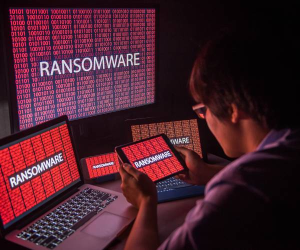 a man sitting in front to screens with computer code and the word ransomware written on one of them