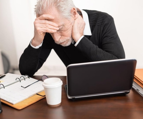 older man with head in hands while looking at computer and notebook