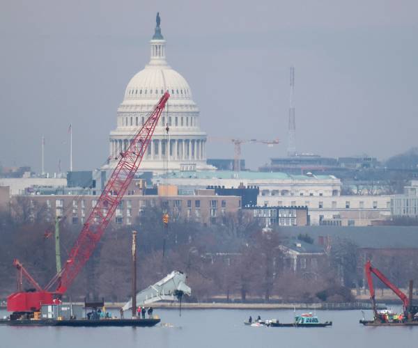 a crane lifts a piece of the airplane with the capitol in the background