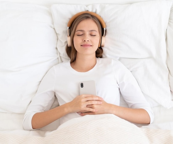 woman in bed with white sheets smiling with headphones plugged into smartphone