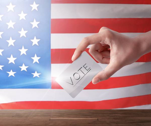 a young-looking hand places a paper that reads "vote" in a box in front of an american flag