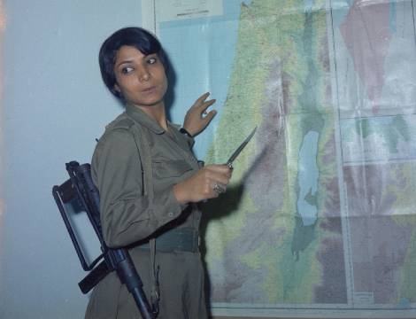 a young khaled in combat fatigues in front of a map of israel and the palestinian territories in with a military rifle