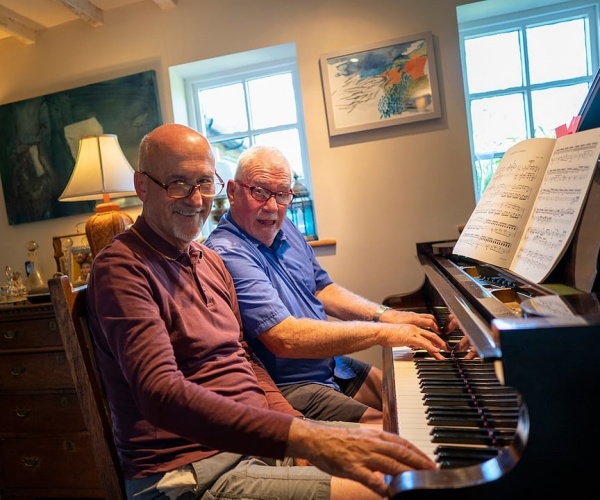 two men sitting at piano smiling, playing music