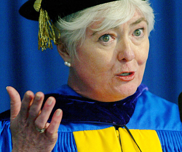 mary beth cahill gestures as she speaks to a graduation ceremony wearing her own cap and gown