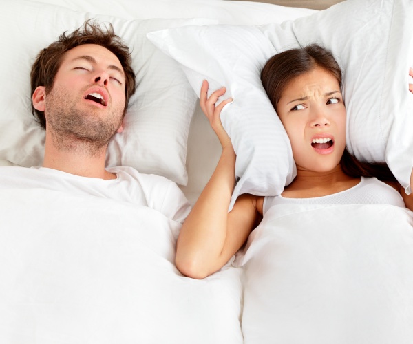 man with sleep apnea sleeping with mouth opened, snoring, while woman puts pillow over her ears