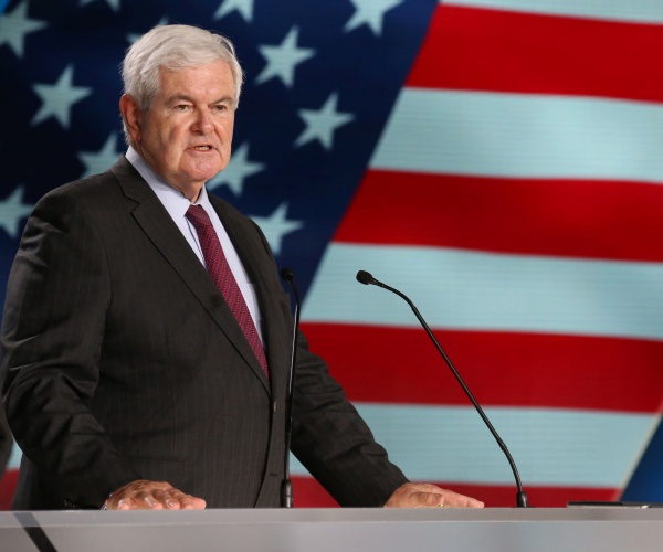 gingrich in a black suit and purple tie with a us flag projection behind him