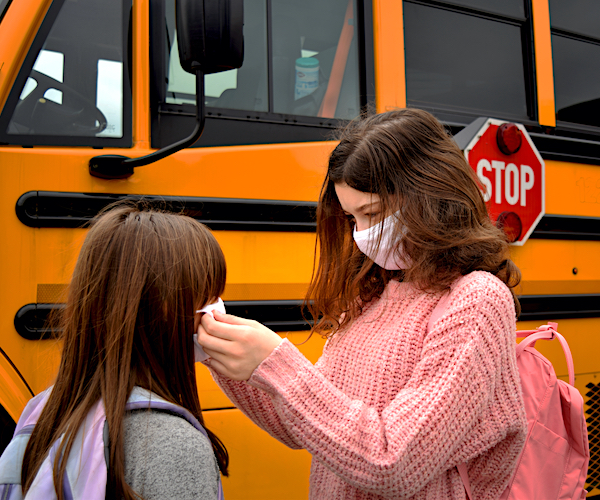 school girls adjust their face masks in front of a yellow school bus