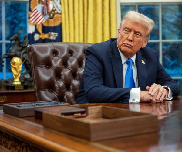 donald trump sits at the resolute desk with his hands clasped on the desk