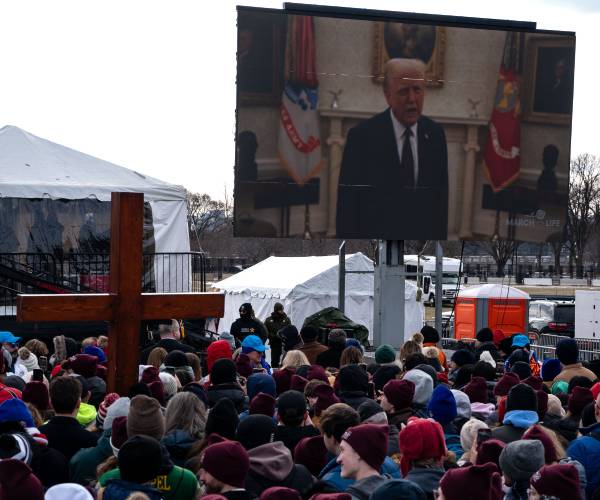 people rallying outdoors in cold weather watch trump on a giant video screen