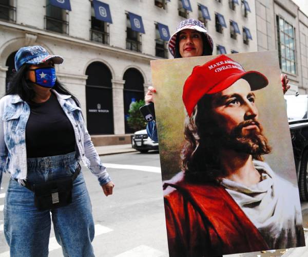 two women standing on the sidewalk with one holding a picture of jesus wearing a make america great again hat