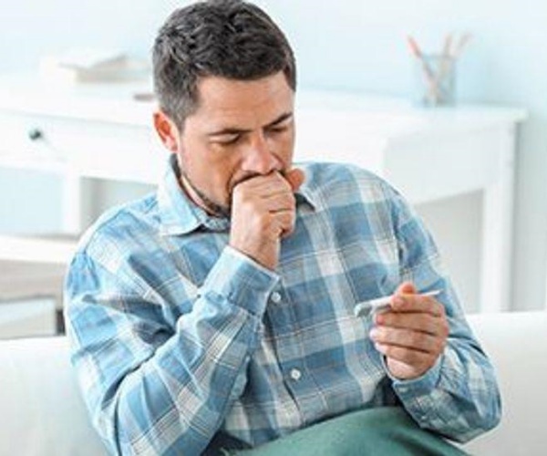 man sitting on couch, coughing and holding thermometer