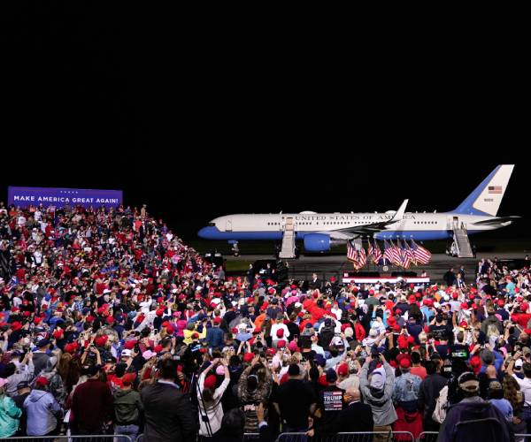 a crowd of people listen to trump during a rally on an airport tarmac with air force one behind him