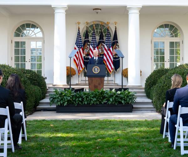 biden standing behind a podium in front of amerian flags with people watching from folding chairs