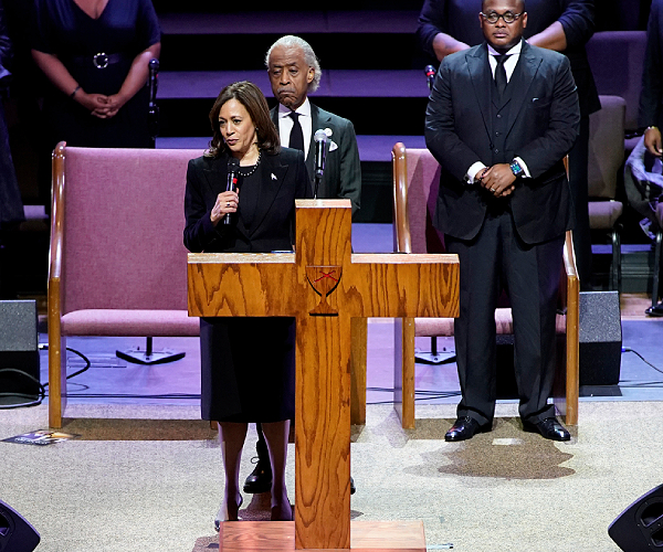 kamala harris stands behind a cross shaped lecturn and speaks into a microphone with the reverend al sharpton behind her