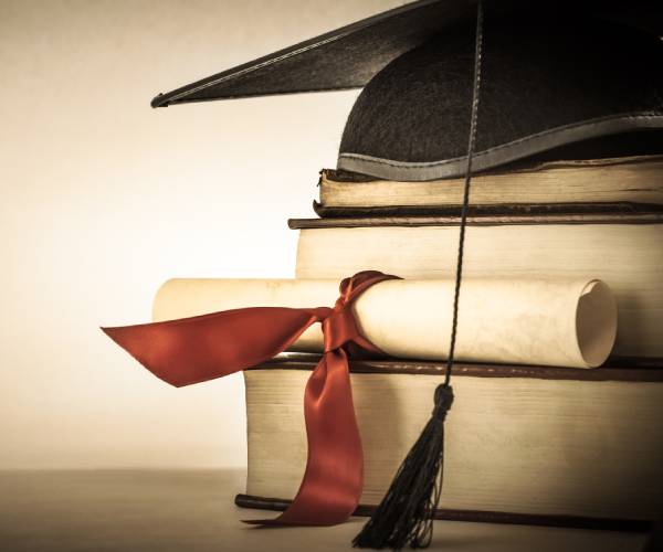 a mortarboard and graduation scroll tied with red ribbon on a stack of old battered books
