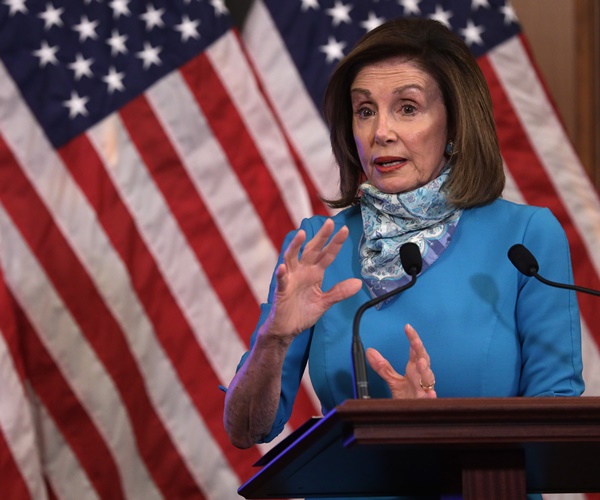 nancy pelosi, wearing a scarf, speaks against a backdrop of u.s. flags