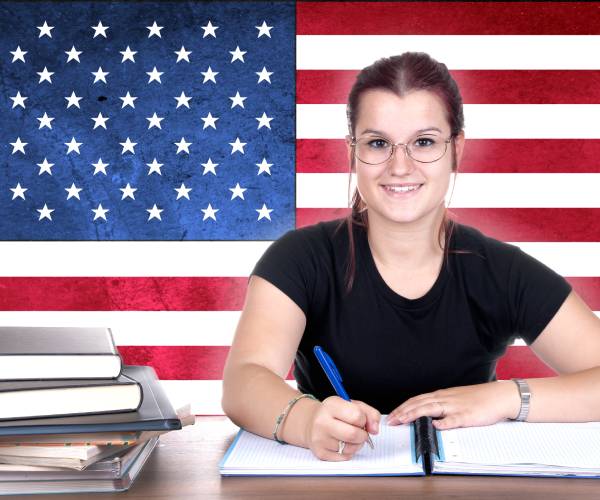 a student doing classwork with a pile of books next to her and an american flag in the background