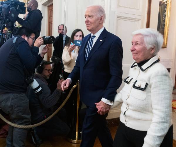 joe biden and a woman hold hands walking into a room of journalists and others