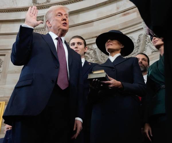 trump with one hand raised and the other at his side while melania holds two bibles beside him