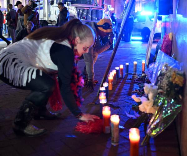 a woman kneels in front of a row of candles and people walk by on the street behind her