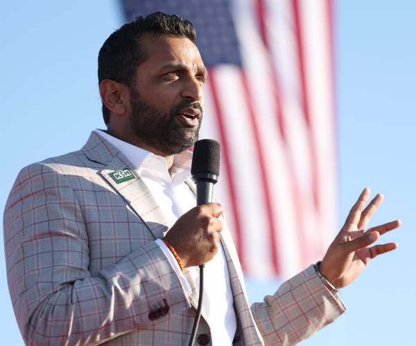 kash patel holds a mic and stands in front of an american flag