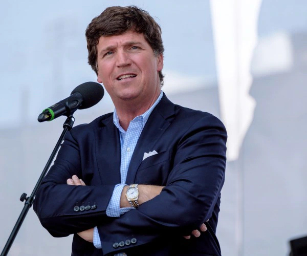 Tucker Carlson speaks into a microphone with his arms crossed in front of him