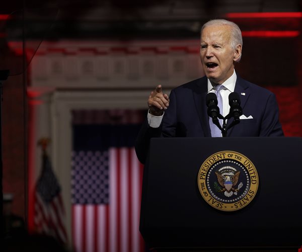 joe biden at a lecturn with a marine and american flag behind him and independence hall drenched in red
