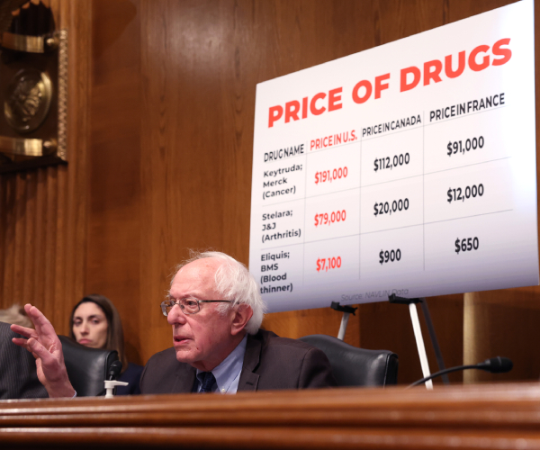 sanders seated with a chart showing drug prices behind him