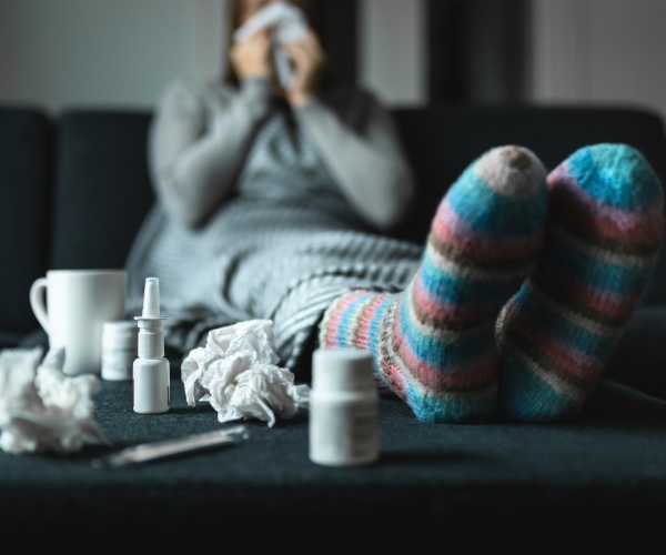 woman sick, sitting with feet up, lots of medicines, blowing nose