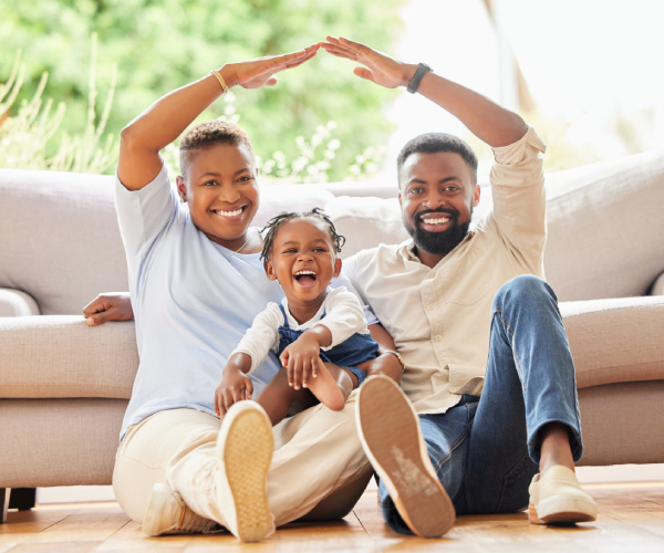 a black family sits in front of a couch and the man and woman make a roof with their arms 