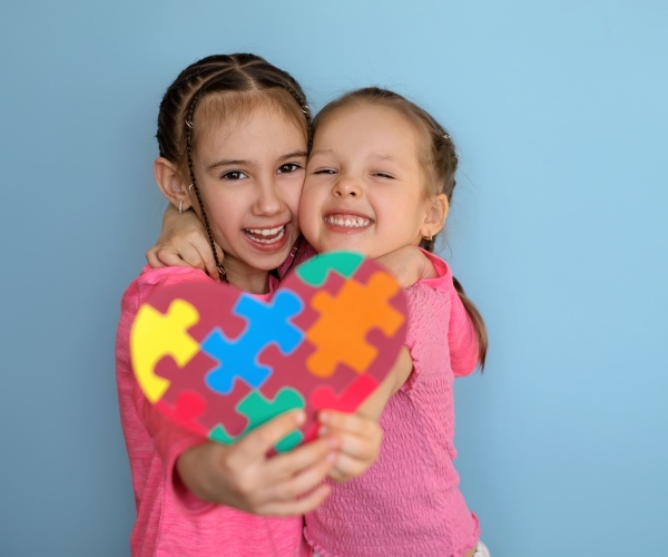 two girls smiling, holding a heart with jigsaw pieces, signifying autism