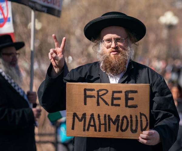 a man in hasidic clothing makes a peace sign with one hand and holds a cardboard sign reading free mahmoud in the other