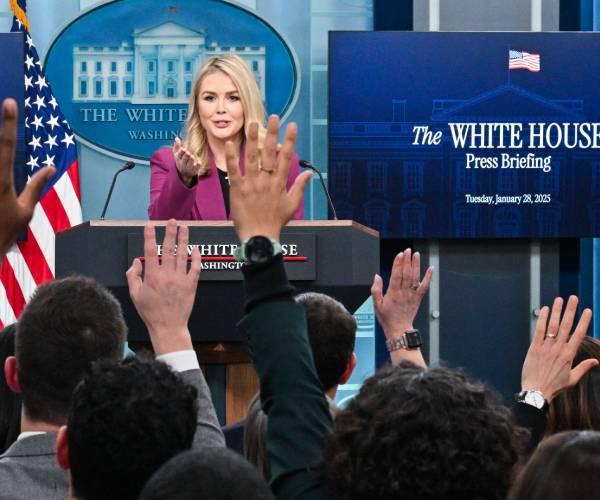 karoline leavitt standing at the podium with the hands of press corps members raised in front of her to ask questions