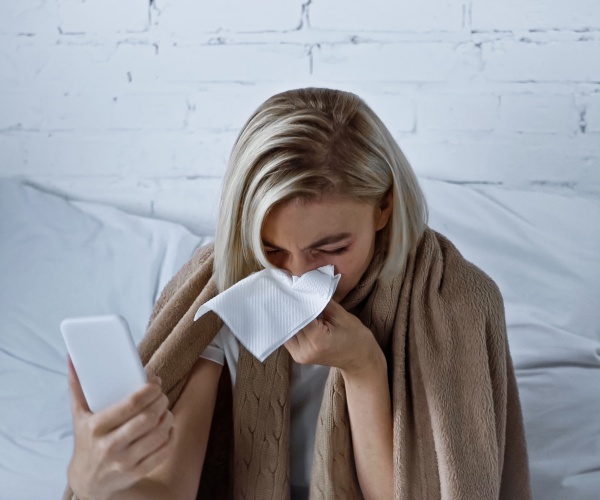 woman holding up phone while sneezing into a napkin